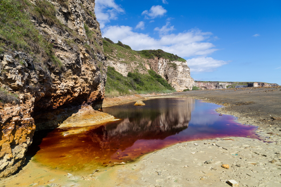 Blast Beach cliffs with a large pool of red and purple water at their foot and blue skies above. County Durham